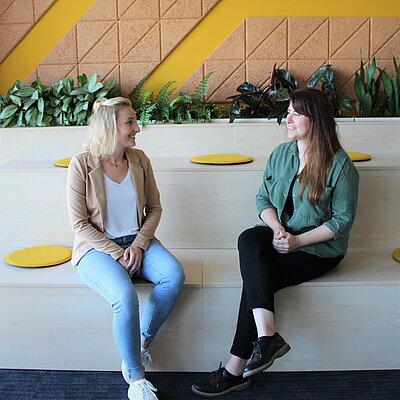 Zwei lächelnde Frauen sitzen zusammen in einer modernen Büroumgebung.