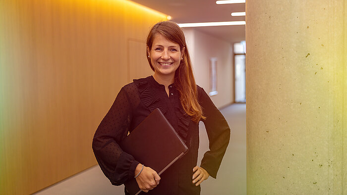 A smiling woman holding a laptop in an office corridor.