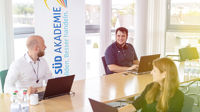 Three people sitting at a table with laptops and talking.