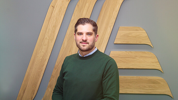 A man in a green jumper standing in front of a wooden wall.