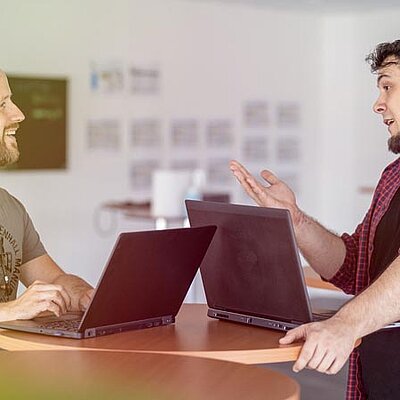 Zwei Männer unterhalten sich an einem Tisch mit Laptops.