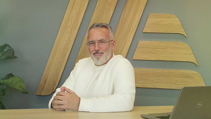 A bearded man in a white jumper sitting at a desk with a laptop.