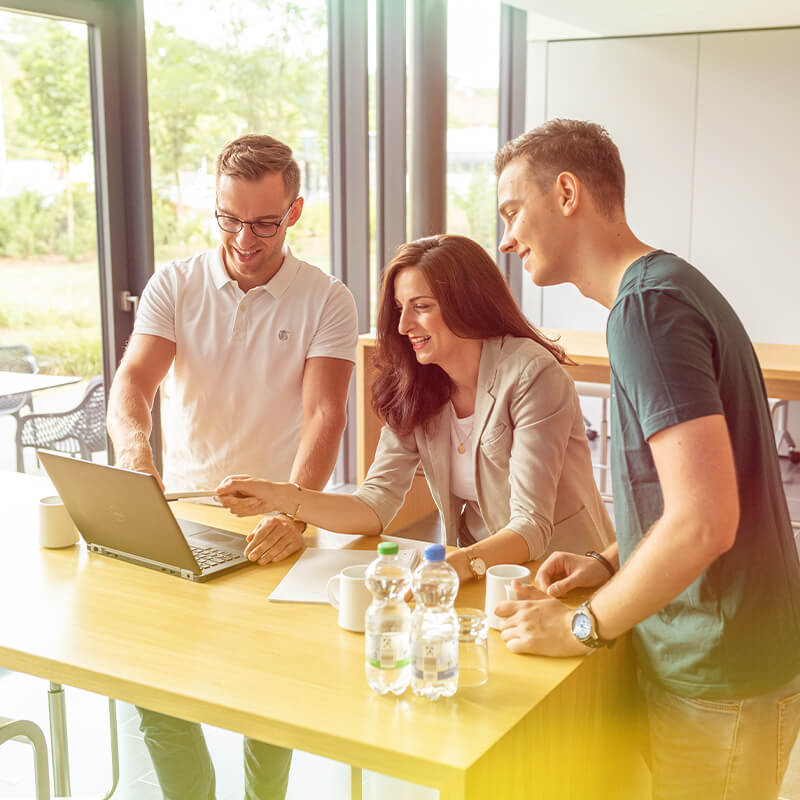 Three people standing around a table with a laptop. They seem to be having a meeting.