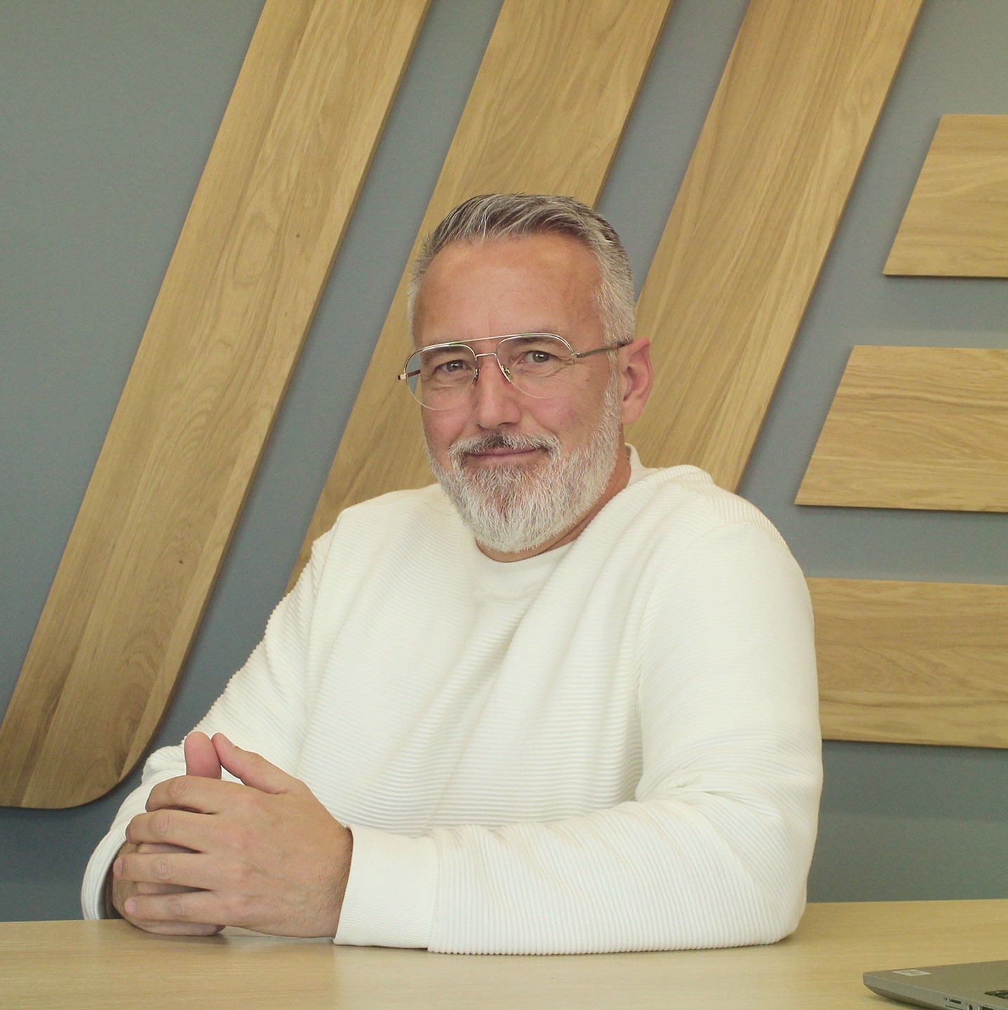 A bearded man in a white jumper sitting at a desk with a laptop.