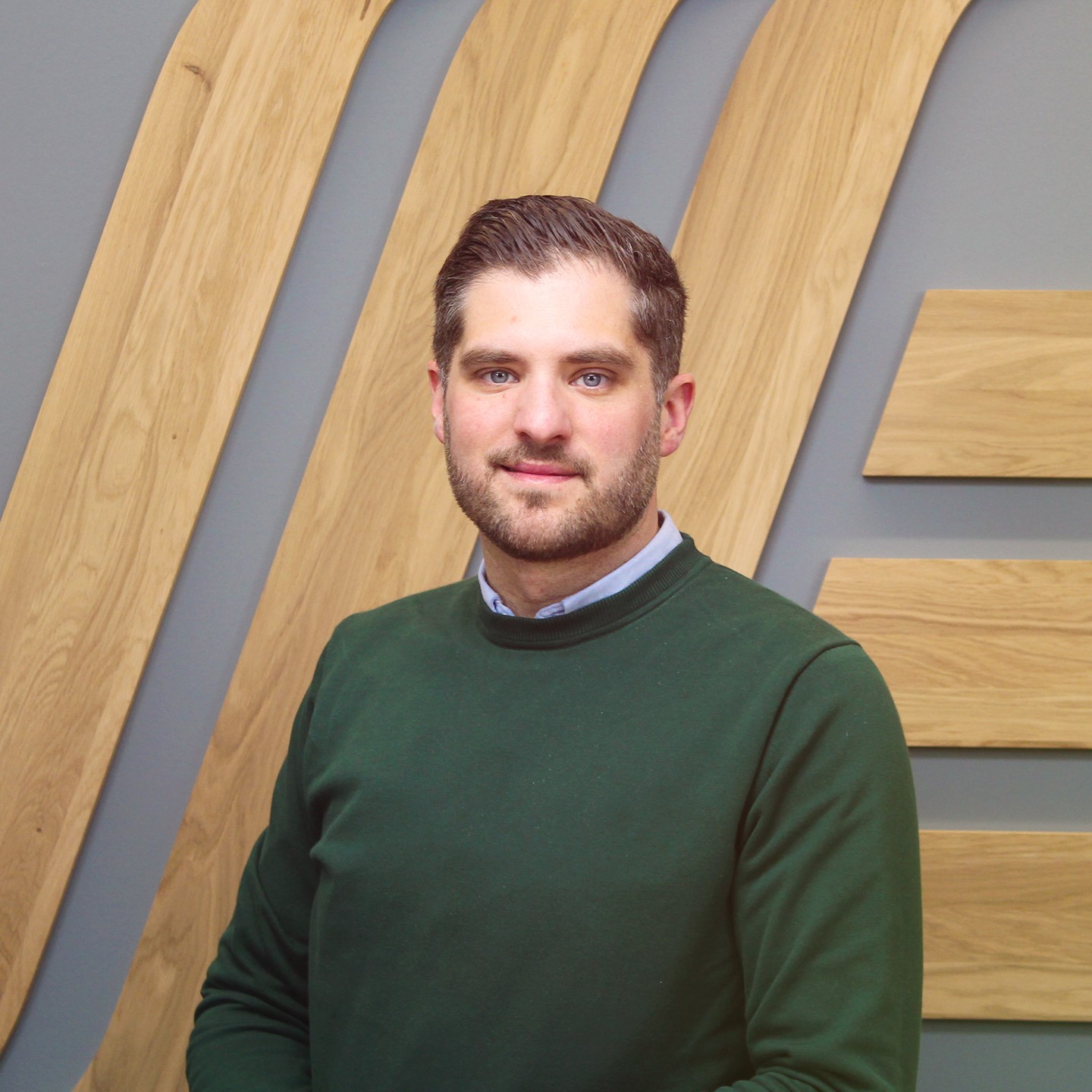 A man in a green jumper standing in front of a wooden wall.