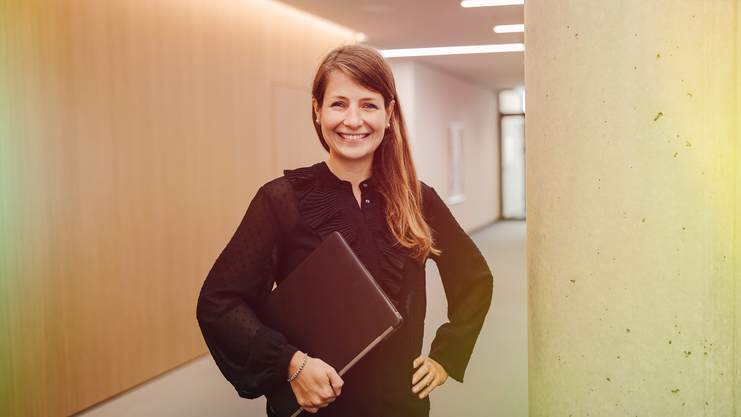 A smiling woman standing in an office with a laptop in her hand.