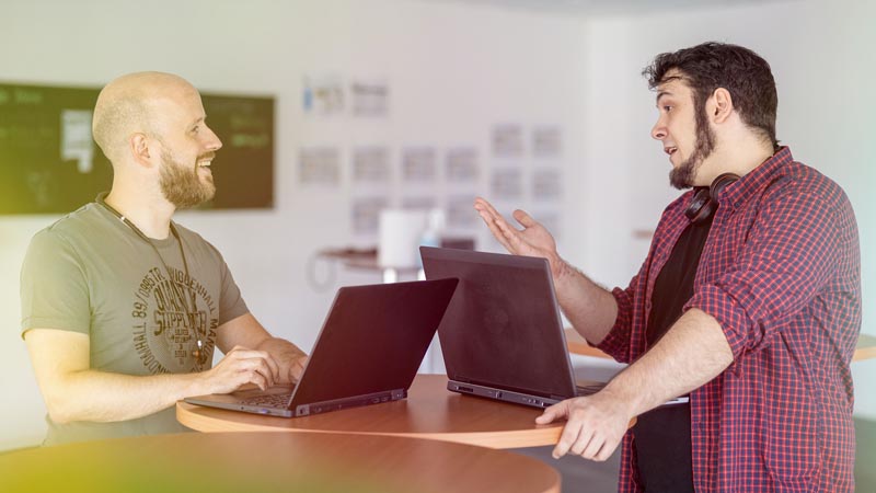 Two men talking at a table with laptops.